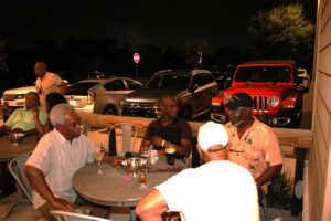 A group of people sitting at a table in front of some cars.